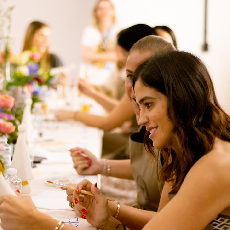 Participantes pintando com pincel em mesa decorada com flores, em workshop de pintura